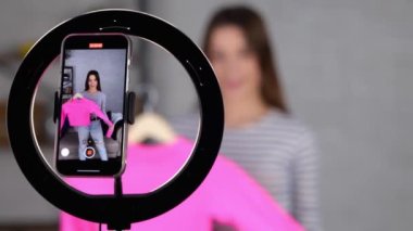 Beyaz tenli güzel bir kadın evinde canlı yayın sırasında çevrimiçi kıyafet satıyor. Genç bayan müşteriye mal gösterirken cep telefonu kullanıyor. Küçük işletme sahibi, online pazar konsepti. - Evet. Gerçek zamanlı.