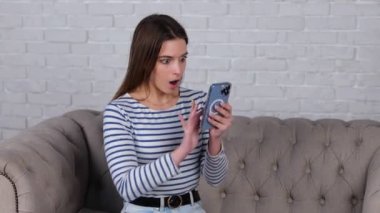 Evdeki bir kanepede otururken akıllı telefondan kötü haberler okuyan Kafkasyalı kadın için endişeleniyorum. Alet kullanırken nahoş haberler okuyan genç bir bayan. Gerçek zamanlı video.