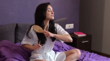 Yatak odasında otururken saçlarını tarayan mutlu kadın. Sabahlık giymiş bir kadın. Sabah rutini. Kişisel bakım prosedürleri kavramı. Gerçek zamanlı video.