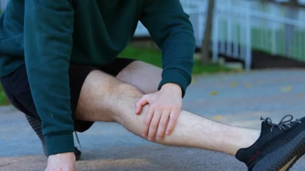 若い白人男性が座って膝 緑の公園の筋肉を伸ばしました 重度の痛みを示している男の顔 リアルタイム スポーツマンは足に強い痛みを抱えている 男は彼の足を暖める — ストック動画