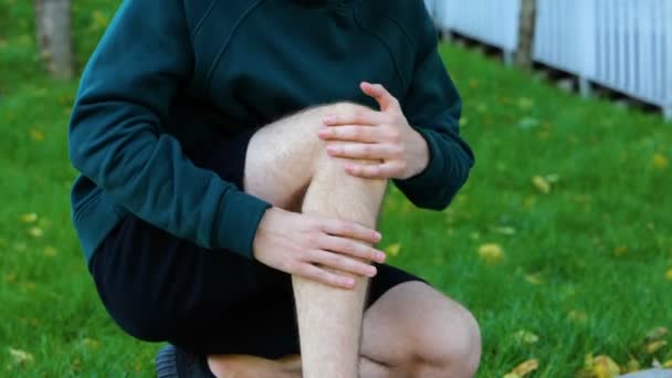 在室外活动前的阳光灿烂的夏日 年轻人在绿地公园按摩他的膝盖 腿和肌肉 运动员穿着黑斑短裤 训练鞋放松 靠拢脚 — 图库视频影像
