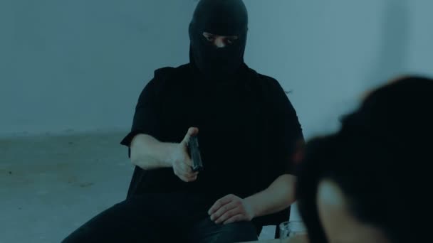 穿着黑色巴拉克拉瓦的男人坐在地下室的桌子旁边用枪威胁女士绑架者喝酒 暴力概念 实时视频 — 图库视频影像