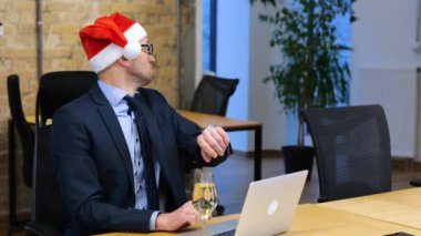 Ofiste tuğla duvarlı bir Noel kutlaması. Beyaz adam bilgisayarında çalışıyor ve saatine bakıyor. Noel Baba 'nın kafasını çıkarıyor ve bir bardak köpüklü şarap içiyor..