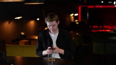 Barda oturan üzgün adam elinde akıllı telefonuyla mesaj okuyor. Viski içen genç adam. Yaşam tarzı konsepti. Gerçek zamanlı..