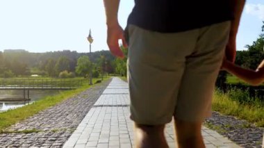 Mutlu bir ailenin yaz günü güzel bir göl kenarında yürüyüşünün arka görüntüsü. Aile zamanı. Gerçek zamanlı. Anne ve baba parkta küçük kızıyla el ele tutuşuyorlar. Anne babasıyla yürüyüşe çıkmış bir çocuk. Doğa