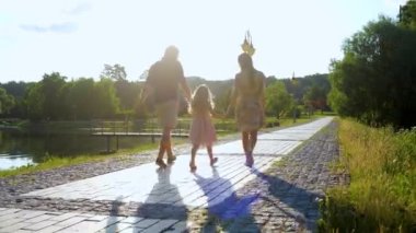 Güneşli bir günde mutlu bir aile birlikte vakit geçiriyor. Anne, baba ve kızı el ele tutuşarak parkta yürüyorlar. Gerçek zamanlı. Mutlu çocukluk kavramı. Aile güzel göl kenarında yürüyor. Arkadan bak. Ebeveynlik