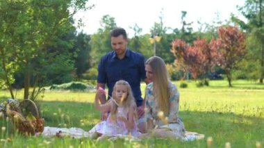 Beyaz bir adam ve kadın, yeşil parktaki çocukla battaniyede oturmuş sabun köpükleriyle oynuyorlar. Mutlu şirin aile yazın dışarıda piknik yapıyor. Aile günü. Çocukluk ve ebeveynlik. Gerçek