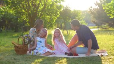 Mutlu bir aile. Genç, neşeli ebeveynler battaniyenin üzerinde oturan çocuklarla doğada vakit geçiriyorlar. Gerçek zamanlı. Yakışıklı baba tatlı kızıyla konuşuyor. Anneye sarılan çocuk. Mutlu bir çocukluk. Ebeveynlik