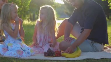 Yazın hafta sonu parkta piknik yapan, battaniyenin üzerinde otururken meyve yiyen beyaz bir aile. Gerçek zamanlı. Mutlu çocuklar piknikte genç ebeveynleriyle dışarıda meyve yerler. Çocukluk