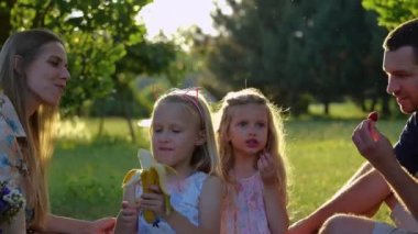 Yazın aileleri parkta pikniğe giden sevimli sarışın küçük kızları kapatın. Beyaz çocuklar dışarıda battaniyenin üzerinde oturmuş meyve yiyorlar. Mutlu bir çocukluk. Aile hafta sonu. Gerçek zamanlı..