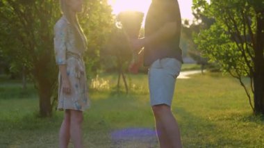 Handsome young man giving beautiful flowers to his pretty happy woman standing outdoor in park on sunset. Caucasian couple in love on nature. Romantic relations concept. Boyfriend and girlfriend