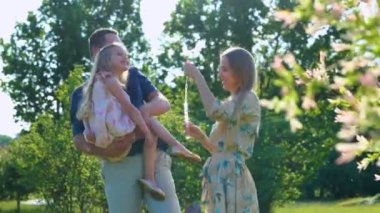 Güneşli bir günde sabun köpüğüyle oynayan mutlu aileye yaklaşan bir kamera. Ebeveynler kızlarıyla doğada oynarlar. Gerçek zamanlı. Çocuk, anne ve babasıyla parkta eğleniyor. Yaz