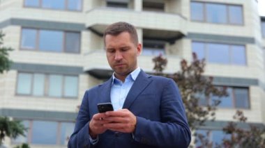 Şehirde ciddi yüzlü beyaz bir erkek müdürün akıllı telefondan mesajlaşırken çekilmiş portresi. Gerçek zamanlı konsept. Genç başarılı iş adamı cep telefonuyla internette geziniyor