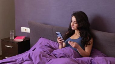 Kredi kartıyla yatakta akıllı telefon kullanarak online alışveriş yapan mutlu bir bayan. Kadın cep telefonu uygulamasında sipariş veriyor, internetten ödüyor. Gerçek zamanlı video.