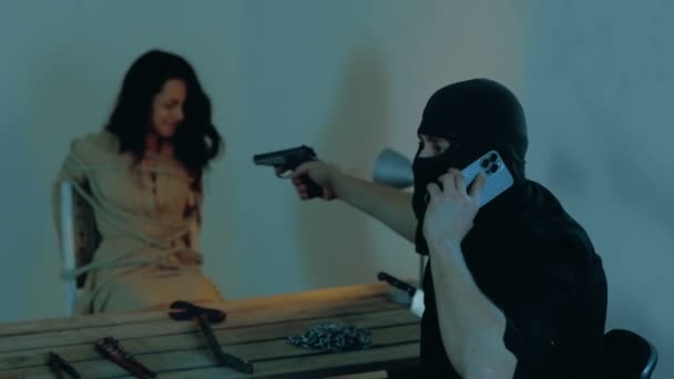 男子绑架者用手机交谈 并在一座废弃的建筑物中威胁一名妇女 穿着黑色巴拉克拉瓦的男人用枪 绑架的概念 实时视频 — 图库视频影像
