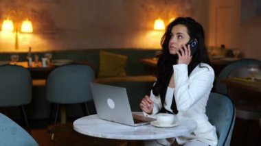 Restoranda dizüstü bilgisayarla çalışırken cep telefonuyla konuşan güzel bir iş kadını. İş, yaşam tarzı konsepti. Gerçek zamanlı video.