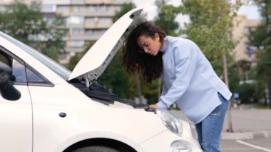Kırık arabanın yanında duran güzel, beyaz, genç bir kadının sorunu çözmeye çalışmasının yan görüntüsü. Gerçek zamanlı video. Kaputu açık aracın yanında bir kadın var. Araba arızası. Araba