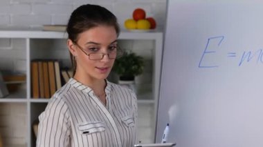 Gözlüklü genç bayan öğretmen evde çalışıyor, online matematik dersi veriyor, tablet tutuyor. Kadın beyaz tahta kullanıyor, cep telefonu tripodda. Çevrimiçi eğitim kavramı. Gerçek zamanlı video.