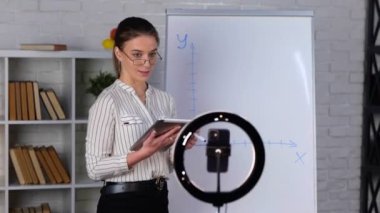 Gözlüklü bayan öğretmen beyaz tahtaya bir grafik çiziyor ve kameraya kayıt dersi veriyor. Çevrimiçi öğrenme kavramı. Gerçek zamanlı video.