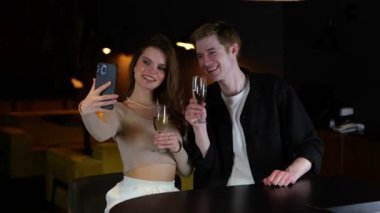 Gülümseyen genç çift restoranda eğlenirken akıllı telefondan selfie çekiyor ve ellerinde şampanya bardakları tutuyorlardı. Yaşam tarzı, ilişki, randevu konsepti. Gerçek zamanlı..