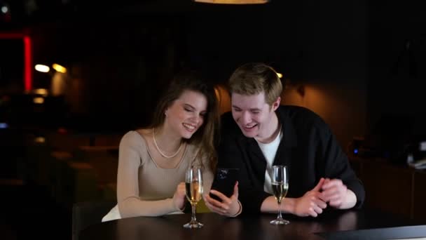 微笑的男人和女人在餐厅休息 一边看智能手机屏幕 一边看新闻 桌上放了几杯香槟生活方式 约会概念 — 图库视频影像