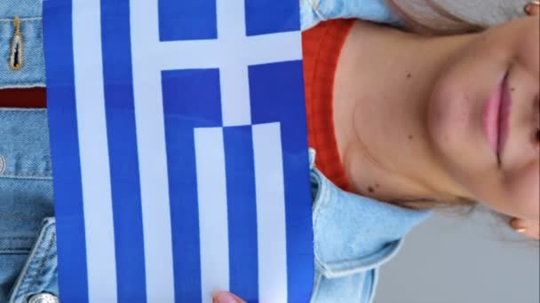 赤いTシャツとデニムのジャケットの美しい十代の少女は 彼女の手にギリシャの小さな旗を保持しています 若い女性は屋外に立って笑顔を浮かべ 小さなギリシャの旗を手で振っている リアル — ストック動画