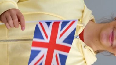 Sarı kapüşonlu İngiliz genç, beyaz, tatlı bayan kameraya gülümseyen Büyük Britanya bayrağı sallıyor. Uzaklaş, yakınlaş, gri arkaplan, kız İngiltere bayrağını elinde tutuyor. Gerçek