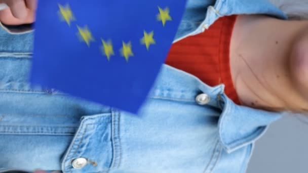 赤のTシャツとデニムのジャケットを着た白人の10代の少女は 彼女の手にヨーロッパ連合の小さな旗を保持しています 美しい若い女性は屋外に立っています 彼女の手で笑顔と欧州連合 の旗を振って — ストック動画