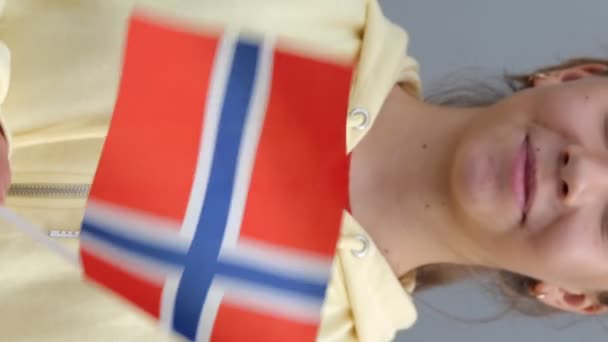 黄色のパーカーの白人の若い美しい10代の少女は カメラで笑みを浮かべて小さなノルウェーのフラグを振っている ズームアウト 閉じる 灰色の背景 小さな女性は彼女の手にノルウェーの旗を維持しています — ストック動画