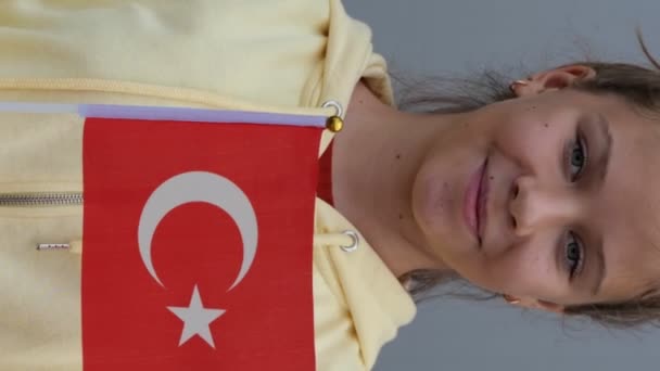 白人の若いトルコの10代の少女トルコの小さな旗を振って カメラで微笑んだ ズームアウト 閉じますアップ グレーの背景 黄色のパーカーの女性の小さな女の子 彼女の手でトルコのフラグを維持 — ストック動画
