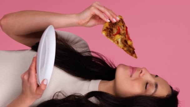 一边站着一边微笑美丽的女性咬着披萨 一边在粉红背景的工作室里摆姿势 生活方式 垃圾食品概念 实时垂直视频 — 图库视频影像