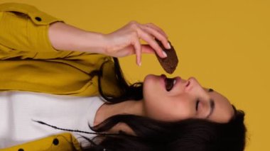 Gülümseyen güzel bir kadın elinde iki yulaflı kurabiye tutarken birini ısırıyor, stüdyoda poz veriyor, sarı arka planda izole halde. Gerçek zamanlı dikey video