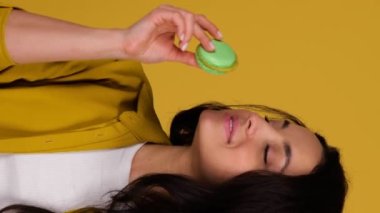 Gülümseyen bayan ısırırken makarondan zevk alıyor. Sarı arka planda duran kadın. Tatlı yemek konsepti. Gerçek zamanlı dikey video