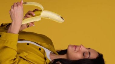 Gülümseyen bayan, sarı arka planda soyulurken muz yiyecek. Sağlıklı, taze gıda konsepti. Gerçek zamanlı dikey video