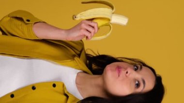 Muz tutarken ayakta durup onu yiyen mutlu güzel bayan. Sarı arka planda izole edilmiş. Sağlıklı, taze gıda konsepti. Gerçek zamanlı dikey video