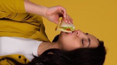 Sandviç yerken stüdyoda poz veren genç bir kadın. Sarı arka planda izole edilmiş. Kopya alanı, insanlar, yaşam tarzı, abur cubur konsepti. Gerçek zamanlı dikey video