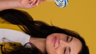 Gülümseyen neşeli genç bir bayan, sarı arka planda izole edilmiş bir şekilde stüdyoda poz verirken lolipopu tutuyor ve onu yalıyor. Gerçek zamanlı dikey video