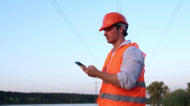 Elektrik mühendisi, akıllı telefon kullanırken güç hattını kontrol ediyor. En yüksek voltaj iletimi. Enerji endüstrisi konsepti. Gerçek zamanlı.
