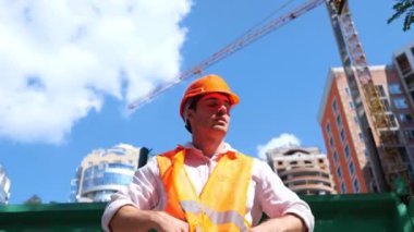 Arka planda inşaat alanı olan açık havada otururken kasket takan kızgın bir erkek işçi. İş, inşaat, endüstri konsepti. Gerçek zamanlı.
