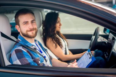 Sınav sırasında gülümseyen erkek eğitmen sınav raporunu tutuyor. Kendine güvenen genç bir kadın araba kullanıyor. Test sürüşü, ulaşım, güvenlik, eğitim kavramı