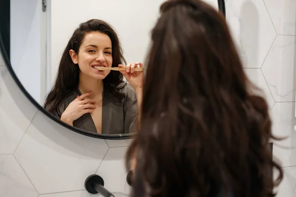 Stock image Back view of woman in grey peignoir standing in the bathroom at home and brushing her teeth. Lady looking in the big mirror. Routine, self-care, lifestyle concept