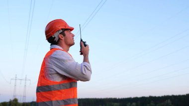 Kasklı genç mühendis dışarıda çalışıyor ve telsiz kullanıyor. En yüksek voltaj iletimi. Enerji endüstrisi konsepti. Yavaş çekim