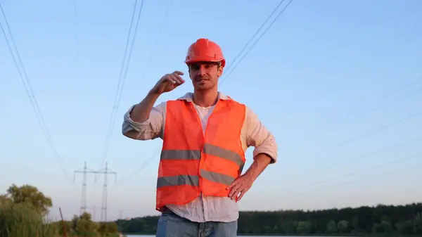 Happy engineer in helmet standing outdoors, high voltage power lines on the background. Highest voltage transmission. Energy industry concept. Slow motion