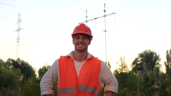 Smiling electrical engineer in helmet posing outdoors in front of a high voltage power lines. Distribution and supply of electricity. Energy industry concept. Slow motion