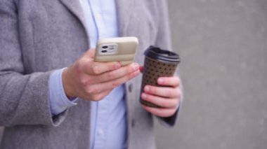 Açık havada duran bir iş adamı akıllı telefonuyla yazarken ve elinde bir fincan kahve tutarken. İş, teknoloji kavramı. Yavaş çekim