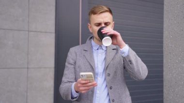 Genç adam kahve içiyor ve binanın yanında dikilirken akıllı telefon kullanıyor. İş, teknoloji kavramı. Yavaş çekim