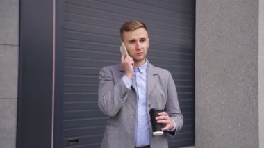 Gülümseyen genç adam cep telefonuyla konuşuyor, kahve içiyor, dışarıda dikiliyor. İş, teknoloji kavramı. Yavaş çekim