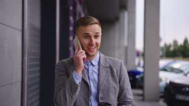 Şehirde yürürken elinde kahve tutarak akıllı telefondan konuşan genç ve mutlu bir işadamı. İş, teknoloji kavramı. Yavaş çekim