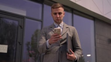 Akıllı telefonda yazarken dizüstü bilgisayar tutan genç bir işadamı, dışarıda duruyor. İş, teknoloji kavramı. Yavaş çekim