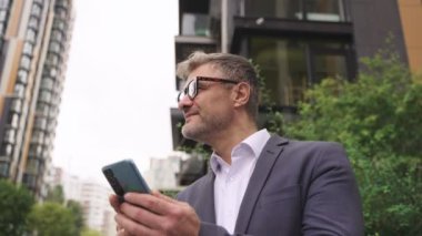 Şehirde akıllı telefon kullanan gözlüklü mutlu bir iş adamı. İş, iletişim, teknoloji konsepti. Yavaş çekim